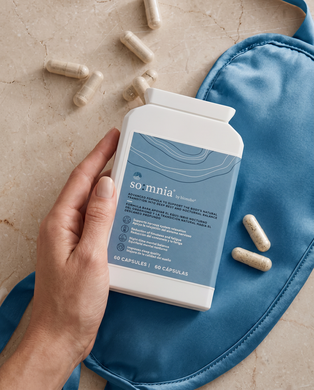 A hand rests a white so:mnia bottle with its dusty blue label on a folded blue silk sleep mask, with pale speckled capsules scattered across the marble surface around it.